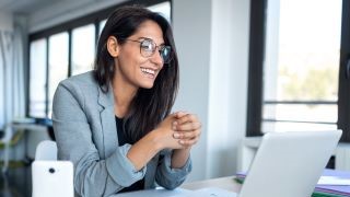 A smiling woman in professional attire sitting in front of an open laptop.