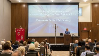 Catechesis News Story 2022 320x180 Staff members sitting in chairs as Dianne Traflet delivered the opening and closing remarks, with a presentation that says The Joy of Discovering and Communicating Our New Life in Christ: Catchesis in the 21st Century