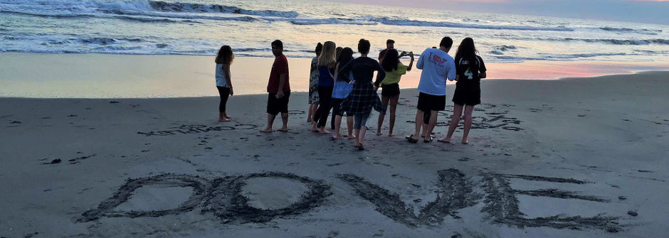 Students on the beach with the word "DOVE" spelled out in the sand. 