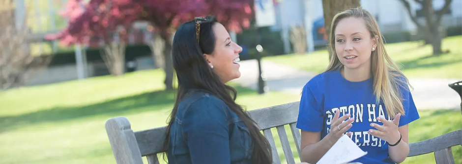 Image of two students talking with books on a bench outside 
