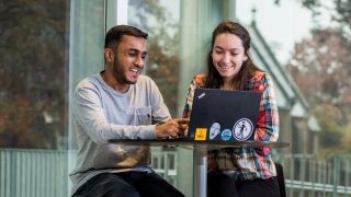 Two smiling Seton Hall students sitting at a high top table looking at an open laptop screen.