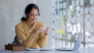 A woman with headphones on playing a game on her phone in front of an open laptop.