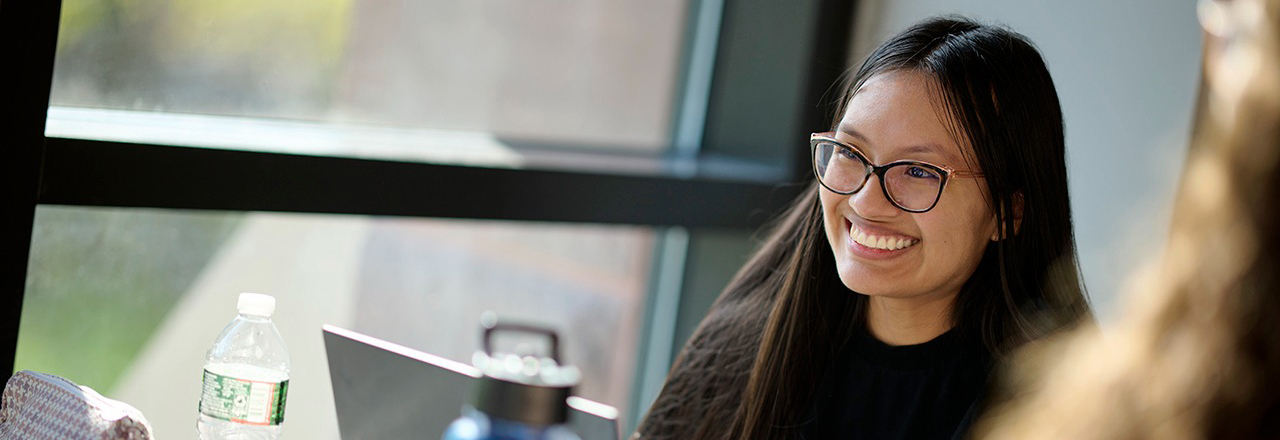 A graduate student smiling sitting near a window with a laptop in class.