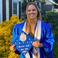 Graduate standing outside of her house in her gown holding her graduation cap. 