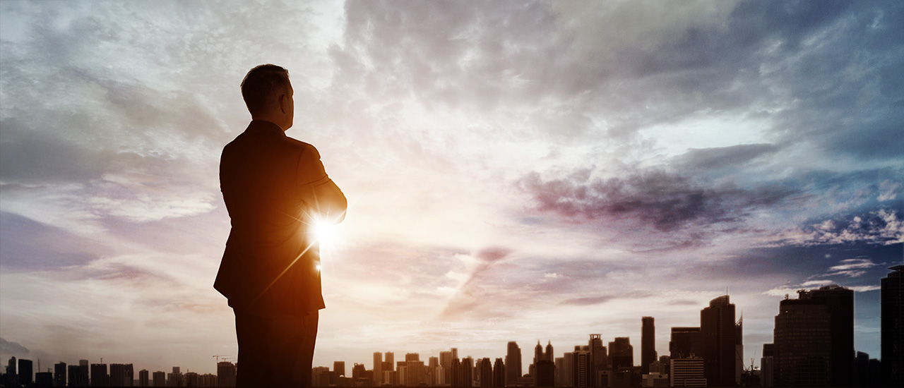 A person in business attire looking out over the city at sunset.