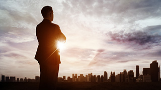 A person in business attire looking out over the city at sunset.