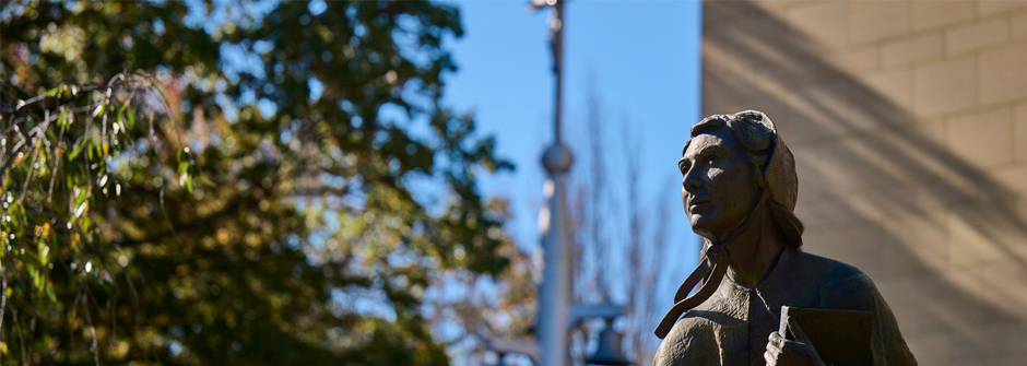 Image of Mother Seton's statue on campus with a cross and a building and a background, next to a tree. 