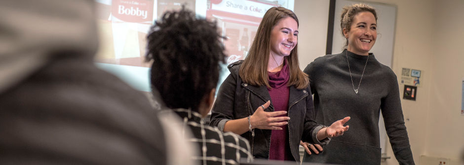 Student presenting at the head of her class, alongside her professor. 