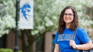 Image of a student wearing a school of business shirt with a backpack standing on campus outside and smiling. 