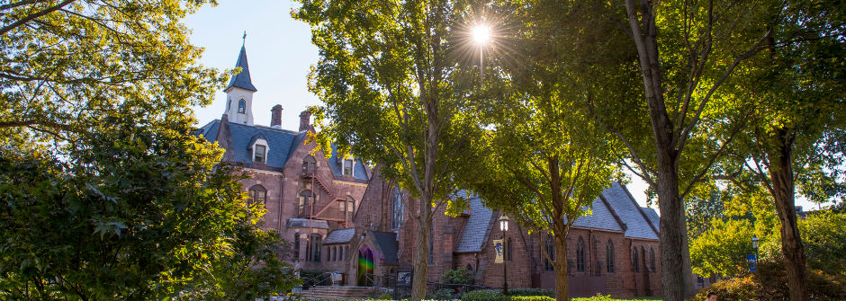An image of President's Hall and the sun shining through trees above the chapel.